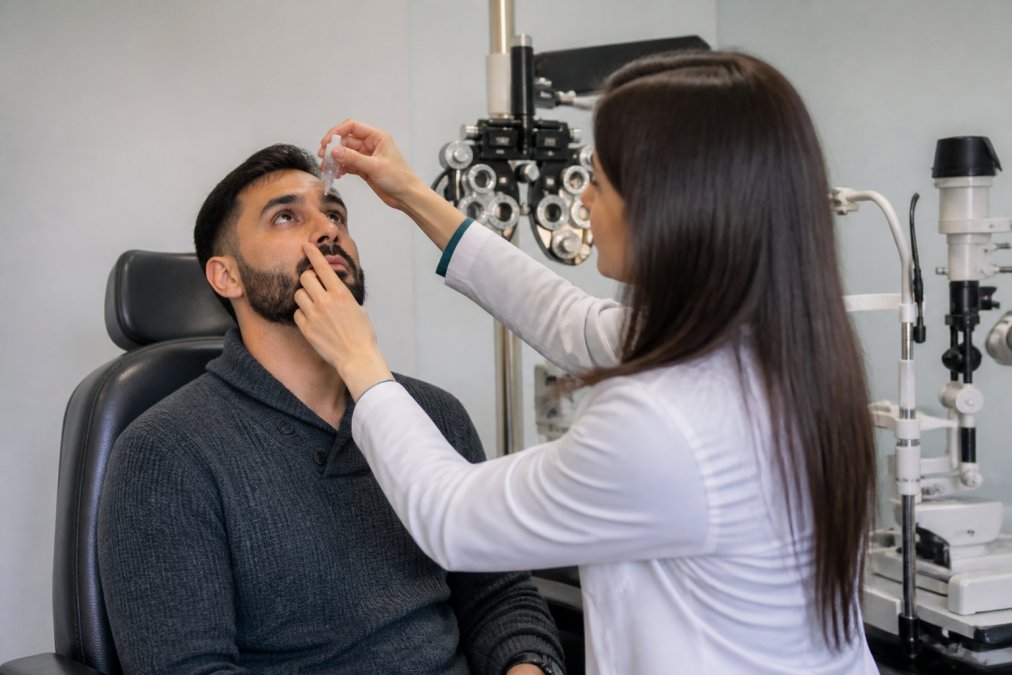 Patient sitting in an optometry exam chair while an eye doctor administers dilating drops, with a phoropter and slit lamp visible in a bright, modern clinic room