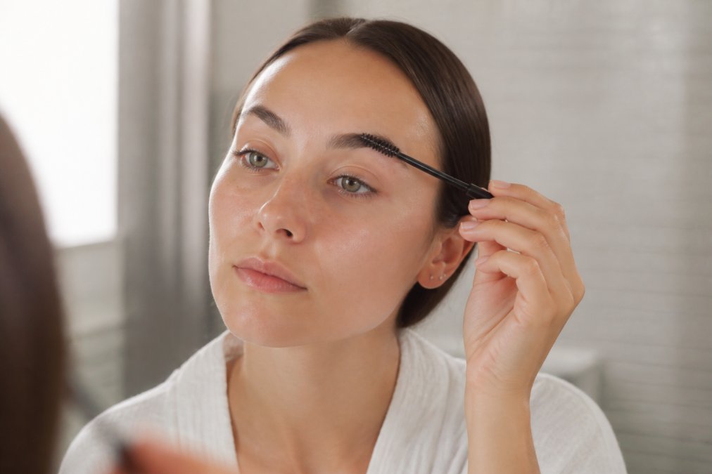 Close-up of a woman gently brushing her laminated eyebrows with a clean spoolie in a bright bathroom, natural daylight, soft neutral tones