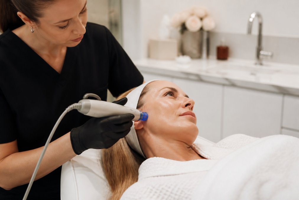 A woman in her 40s receiving a professional collagen-stimulating facial treatment in a modern medspa, with a licensed aesthetician using a handheld device on her cheek while she rests comfortably under soft clinical lighting