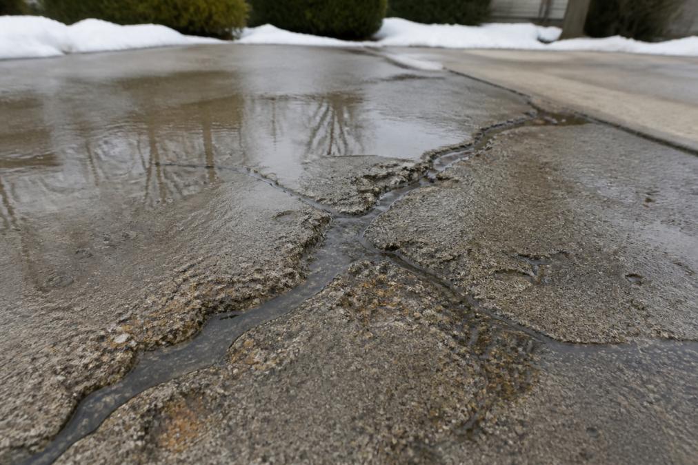 Close-up of concrete driveway showing water pooling in cracks during rainy conditions, with visible frost damage and surface deterioration from freeze-thaw cycles