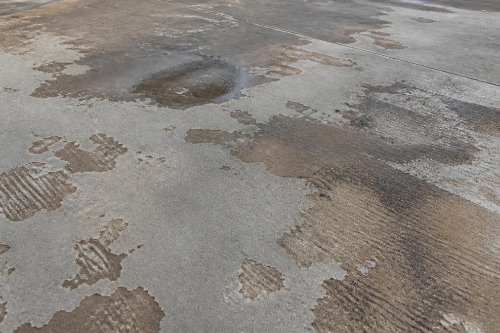 Close-up of a concrete driveway surface showing visible peeling sealer, discoloured patches where water has pooled, and worn traffic marks from vehicle tyres