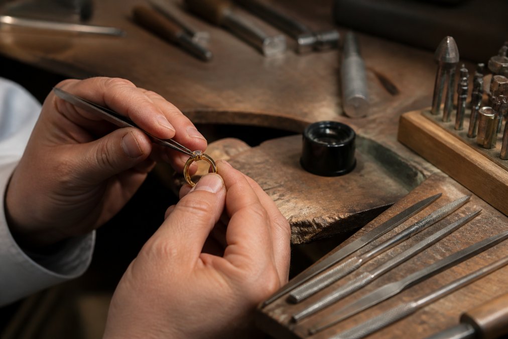 A jeweller's hands using fine tools to set a small diamond into a gold ring on a workbench, with ring files and a loupe nearby in a warmly lit workshop