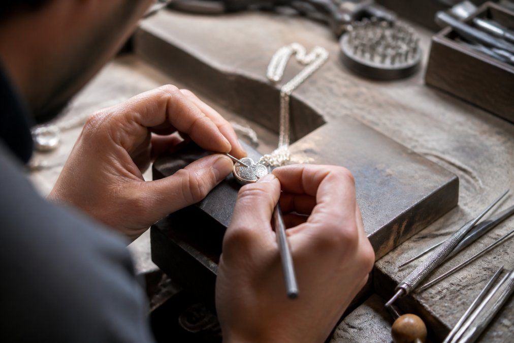 Close-up of a jeweler's hands working on a custom necklace at a workbench, with engraving tools and the piece positioned for detail work, natural light from overhead lamp