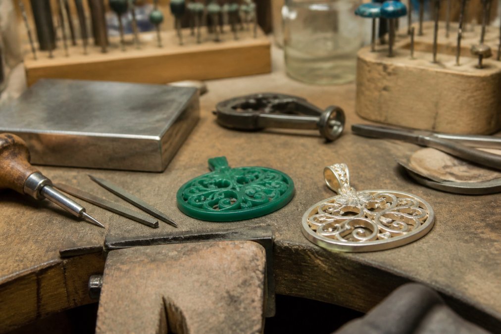 A jeweler's workbench with hand tools, a wax model, and a partially finished pendant under warm workshop lighting