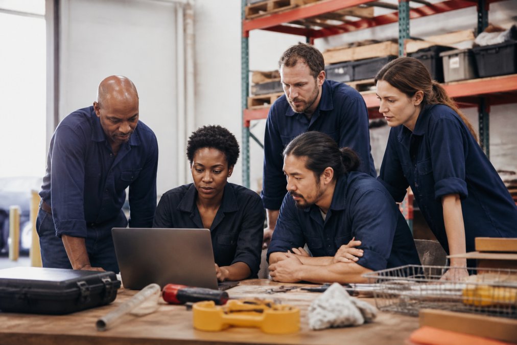 A diverse team of field workers and supervisors gathered around a laptop in a training session, examining the digital work order system with focused attention, natural indoor lighting