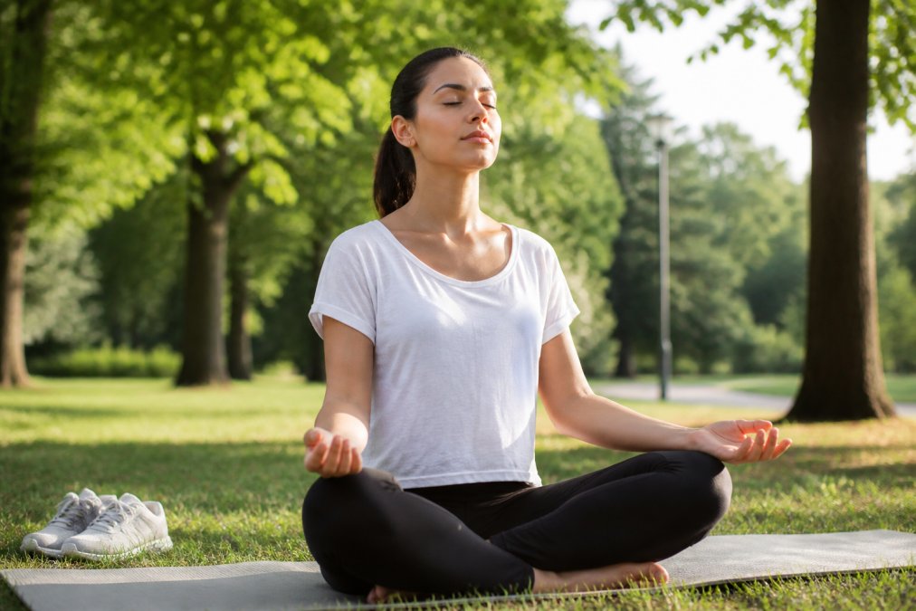 Person sitting peacefully outdoors with eyes closed in natural daylight, practicing mindfulness in a calm park setting with soft morning light filtering through trees