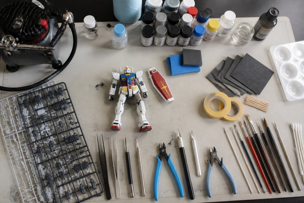 Overhead view of a well-organized painting station with airbrush, spray cans, hand brushes, sanding tools, masking supplies, and a Gundam model kit laid out on a clean work surface