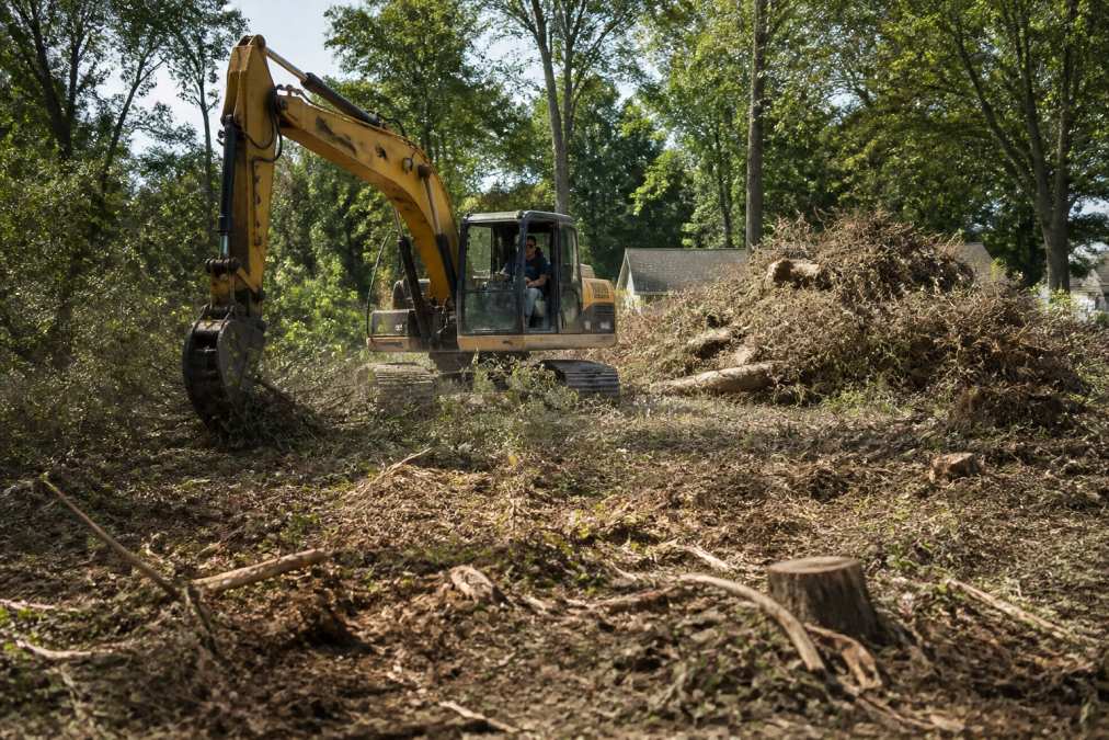 Heavy equipment operator in an excavator clearing thick brush and vegetation from an overgrown residential lot, with large piles of cleared debris and tree stumps visible in the background