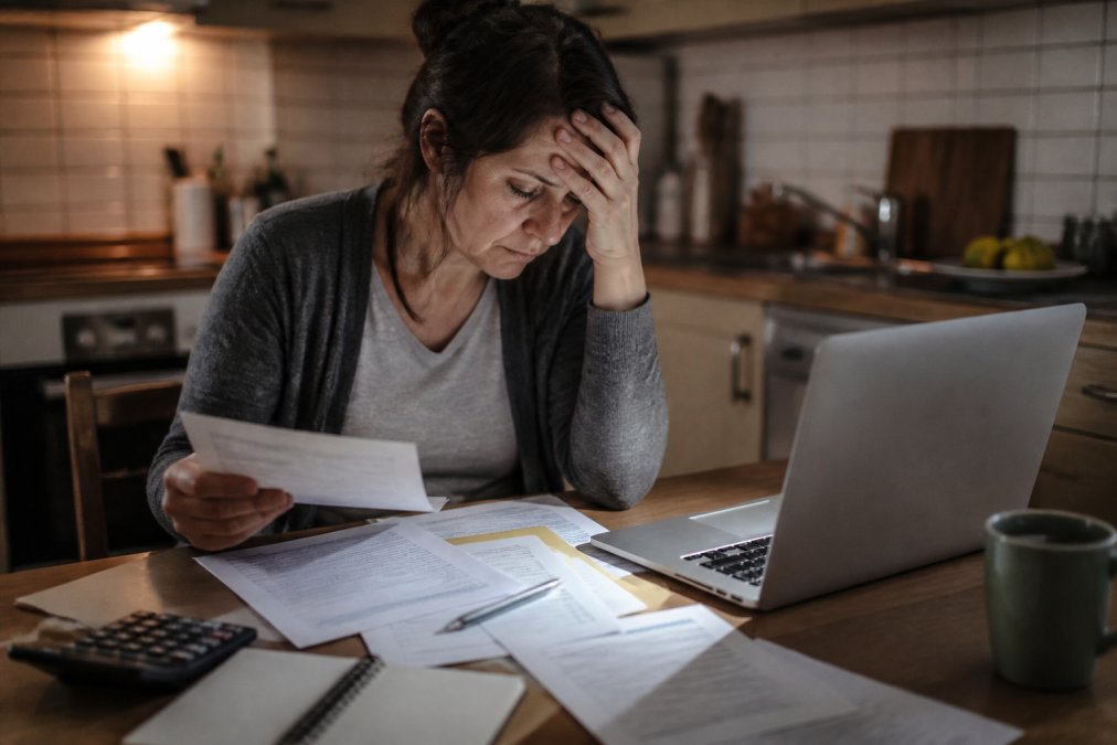 A stressed individual sitting at a kitchen table with medical bills and legal paperwork spread out, looking at a laptop screen under dim kitchen lighting