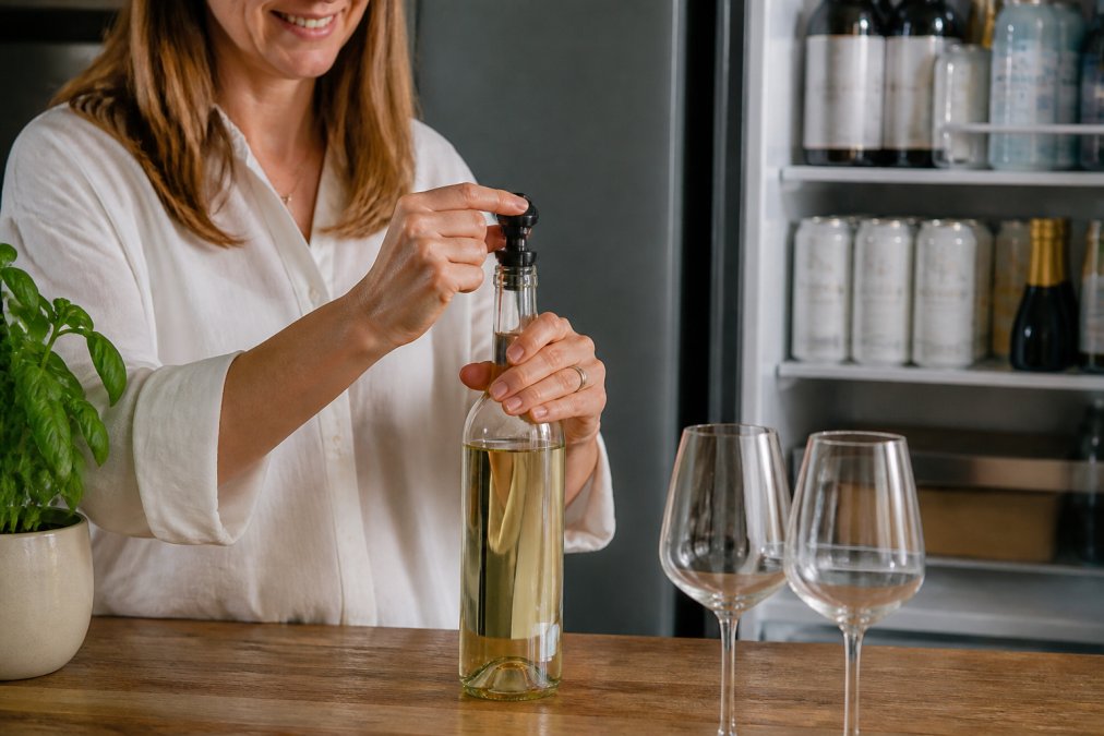 A woman's hands twisting a vacuum stopper into the neck of an opened bottle of alcohol free white wine on a kitchen worktop, a fridge door open behind her with 0% drinks on the shelf