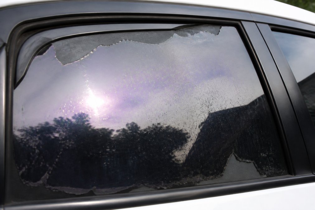 Close-up of a vehicle window showing visible signs of tint deterioration with bubbling near the edges, peeling corners, and purple discoloration across the film surface in natural sunlight