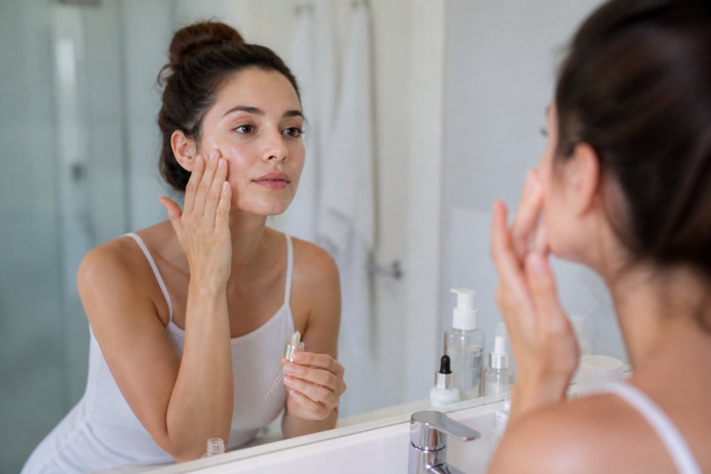 Woman applying retinol serum to her face in a bright bathroom, looking at herself in the mirror with a calm expression, demonstrating proper application technique