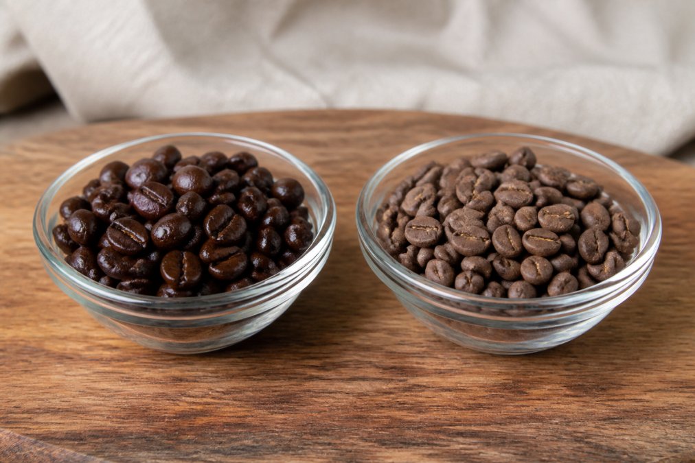Side-by-side of two small glass bowls on a wooden surface, one holding glossy, freshly roasted dark beans with visible surface oils, and the other holding dull, faded, matte older beans under soft natural light