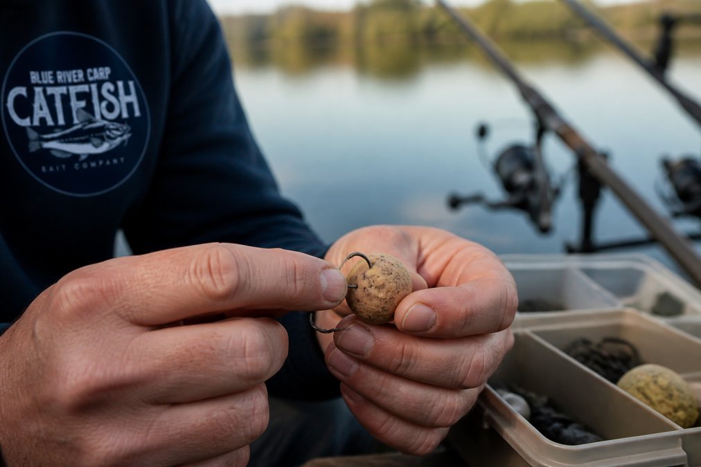 Close-up of a fisherman's hands checking a scent-infused bait ball on a hook above calm water, tackle box visible in background