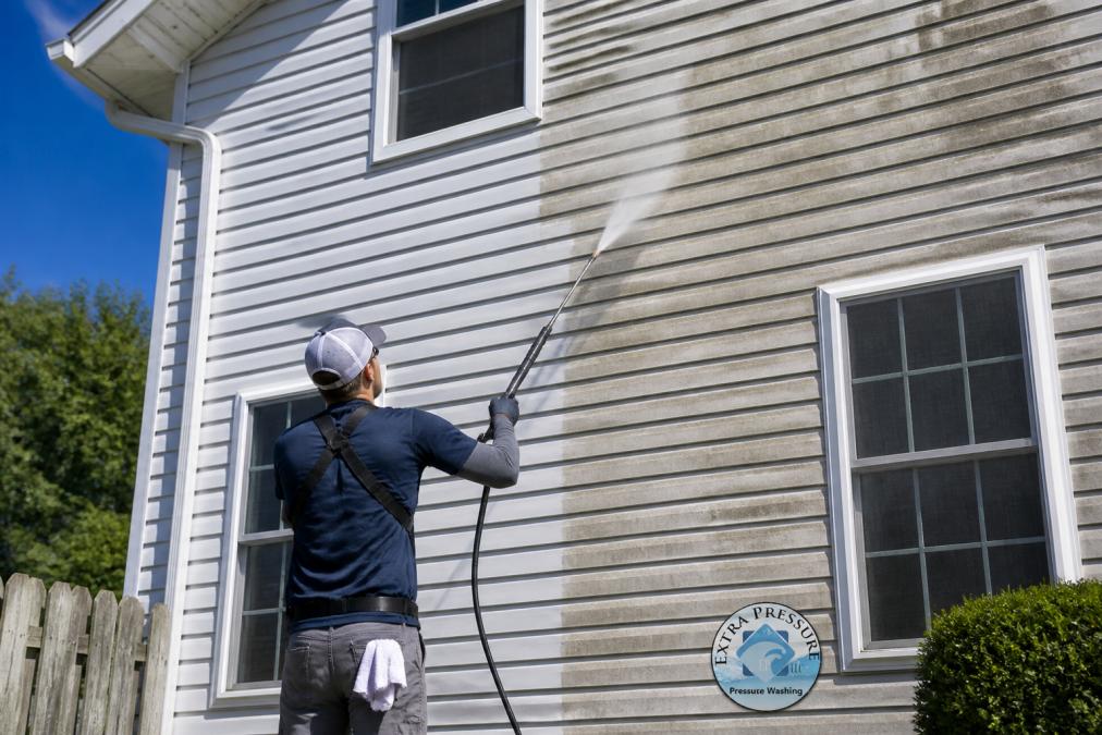 Technician using a soft wash sprayer on vinyl siding to show how long soft washing last on a suburban home