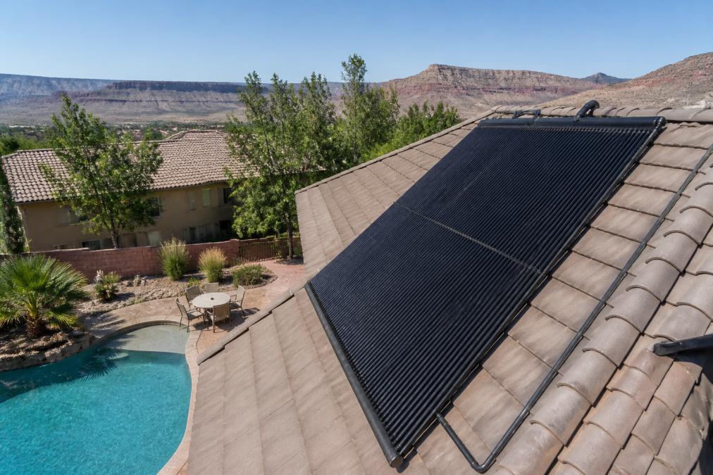 Rooftop view of black solar pool heating panels installed on a residential home, with a blue swimming pool visible below and clear afternoon sunlight