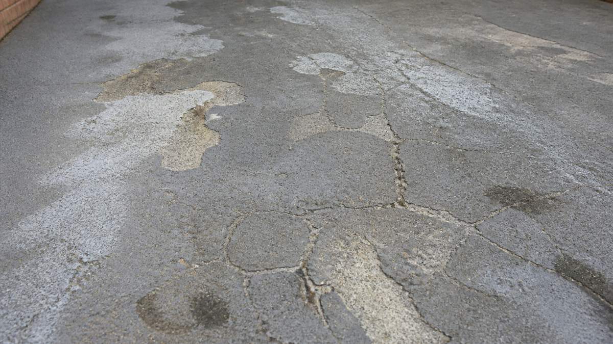Close-up of a deteriorating concrete driveway surface showing visible cracks, peeling coating patches, and discoloured areas in daylight