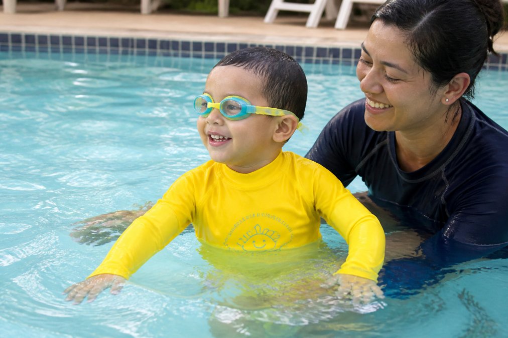 Young toddler wearing a vibrant high-visibility rash guard and swim goggles playing confidently in shallow pool water with an instructor nearby, demonstrating comfort and engagement