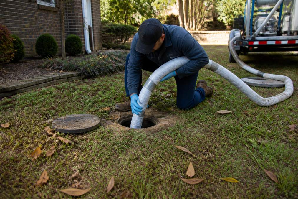 A technician in work gear feeding a resin-impregnated liner into a sewer access point in a residential yard, with minimal ground disturbance visible around the clean-out opening