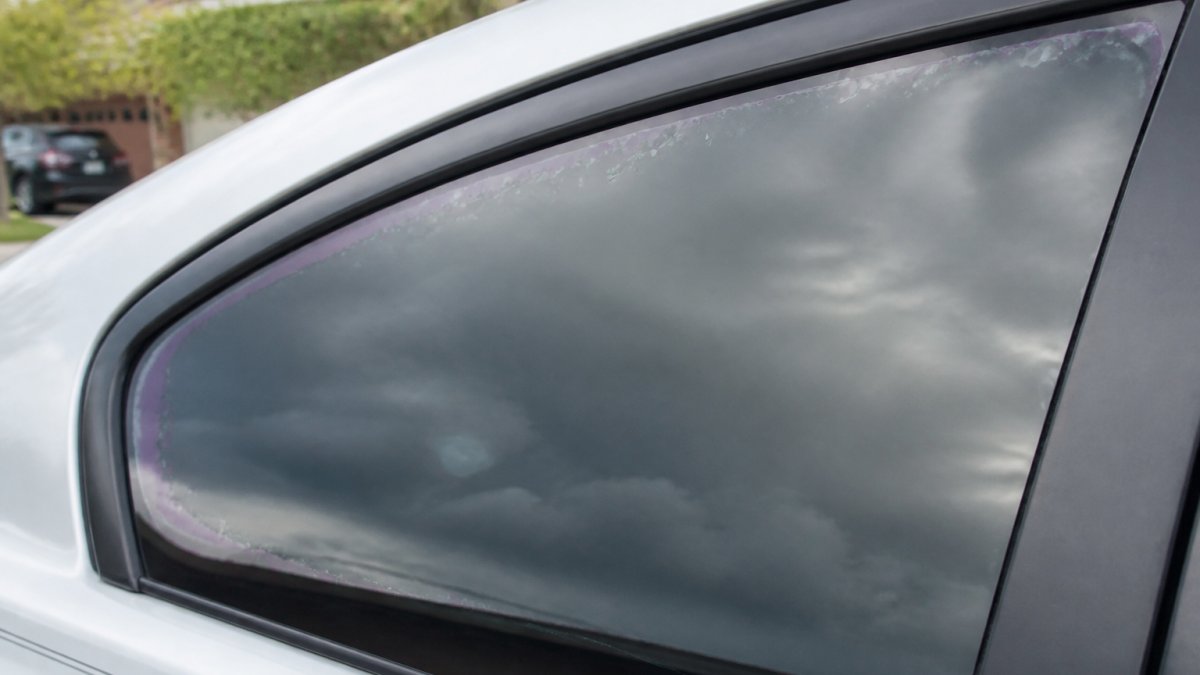 Close-up of a car side window showing early signs of tint aging, with small bubbles and slight purple discoloration visible along the edge, photographed in natural daylight in a driveway