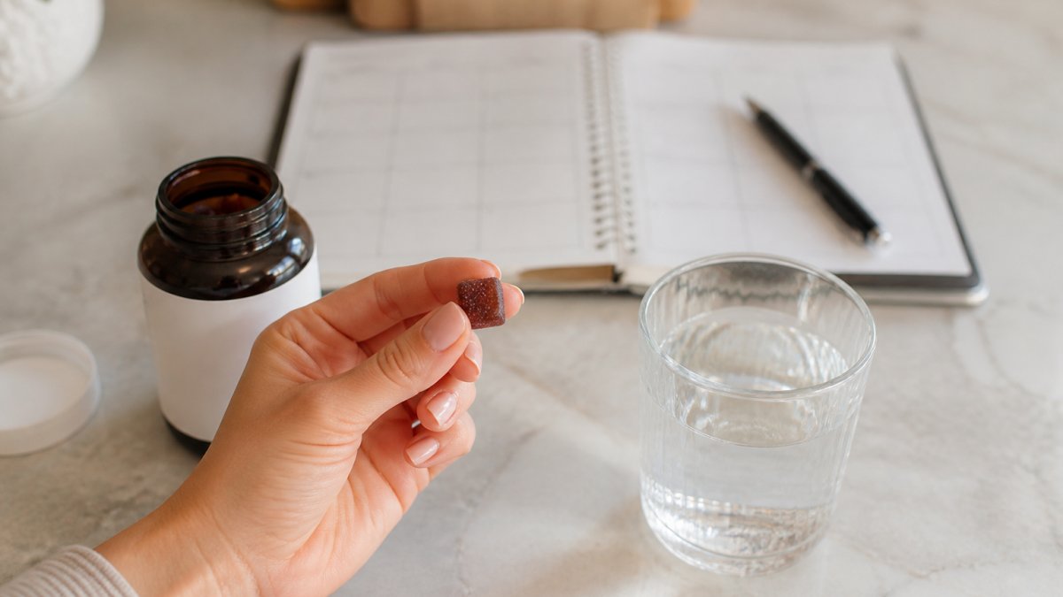 A close-up of a person's hand holding a daily supplement gummy next to a glass of water on a kitchen counter, with a weekly planner and pen in soft focus in the background