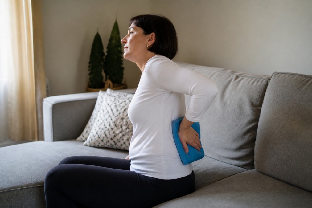 A person applying a heat pack to their lower back while sitting on a sofa at home, natural daylight