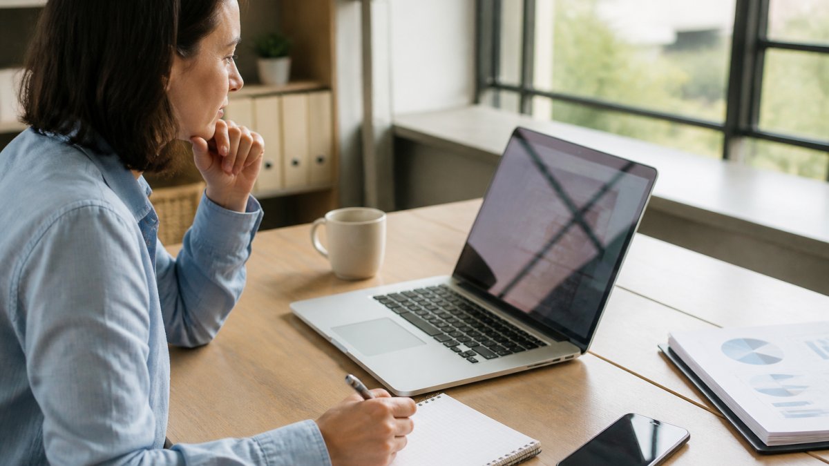 Professional marketer reviewing analytics dashboard on laptop with notebook and pen, tracking SEO performance metrics in bright office with natural daylight