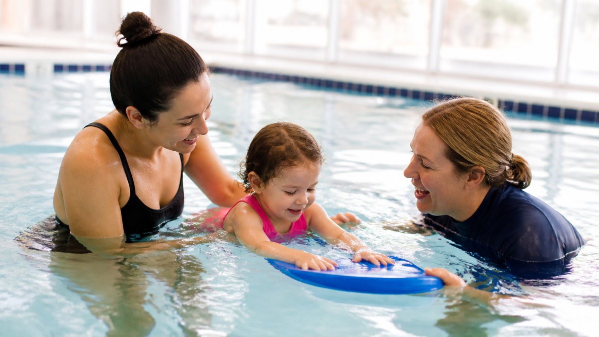 Qualified swim instructor and parent guiding a toddler in a shallow pool during a safety lesson