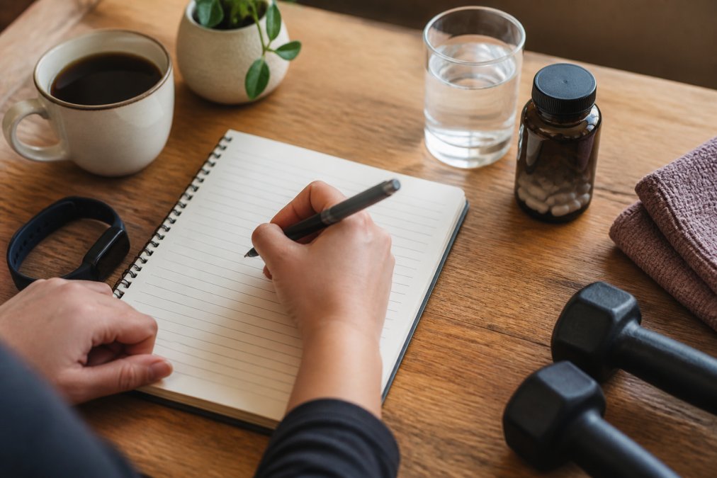 Person writing in a health journal with pen, documenting energy levels and wellness metrics on a desk with morning coffee and fitness tracker