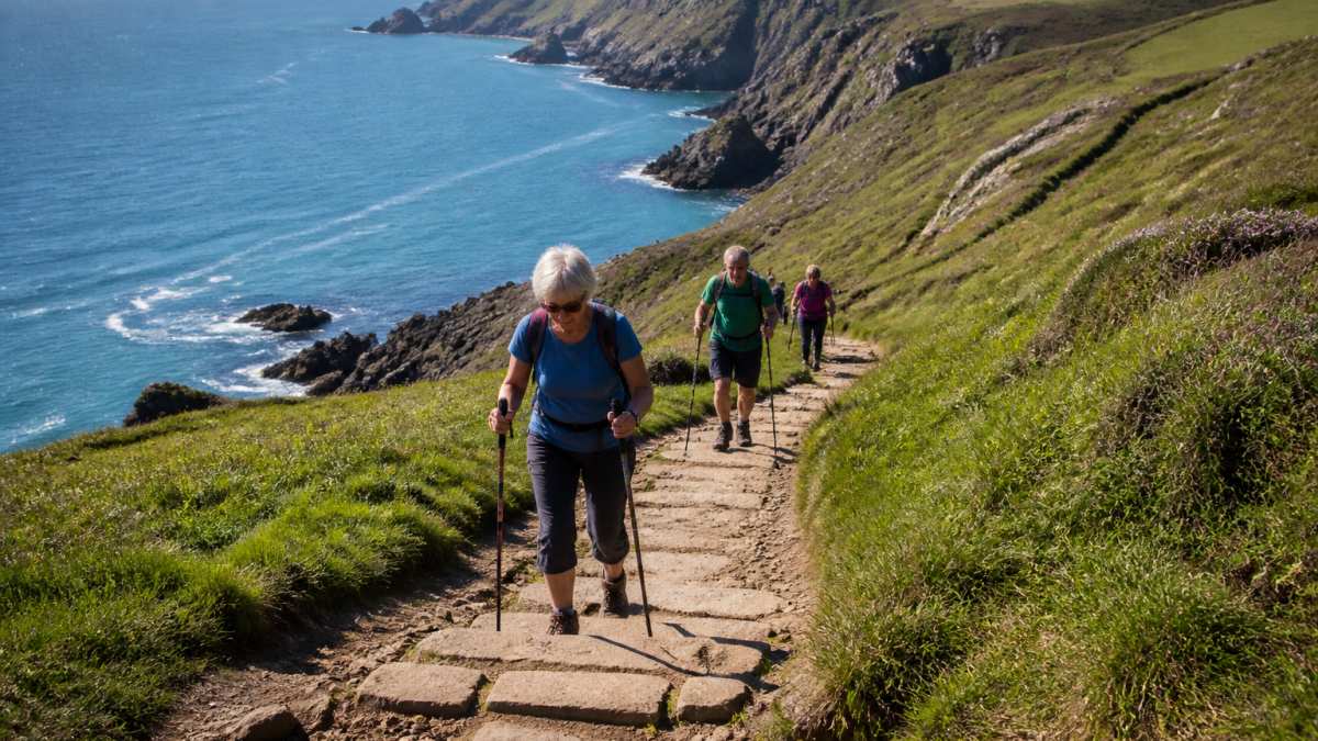 Walkers on a coastal path with dramatic cliffs and sea views in the background, showing varied terrain including grassy sections and stone steps