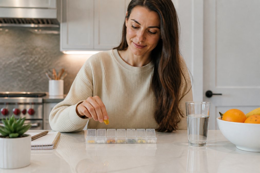 Person organizing daily supplements in a pill organizer on a kitchen counter, with a glass of water and morning routine items visible