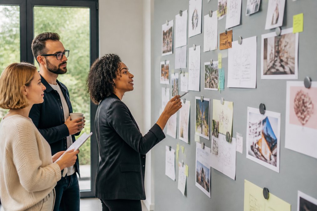 Modern office team collaborating around a magnetic wall displaying project cards, notes, and printed materials held with magnets during a planning session, with natural light from windows