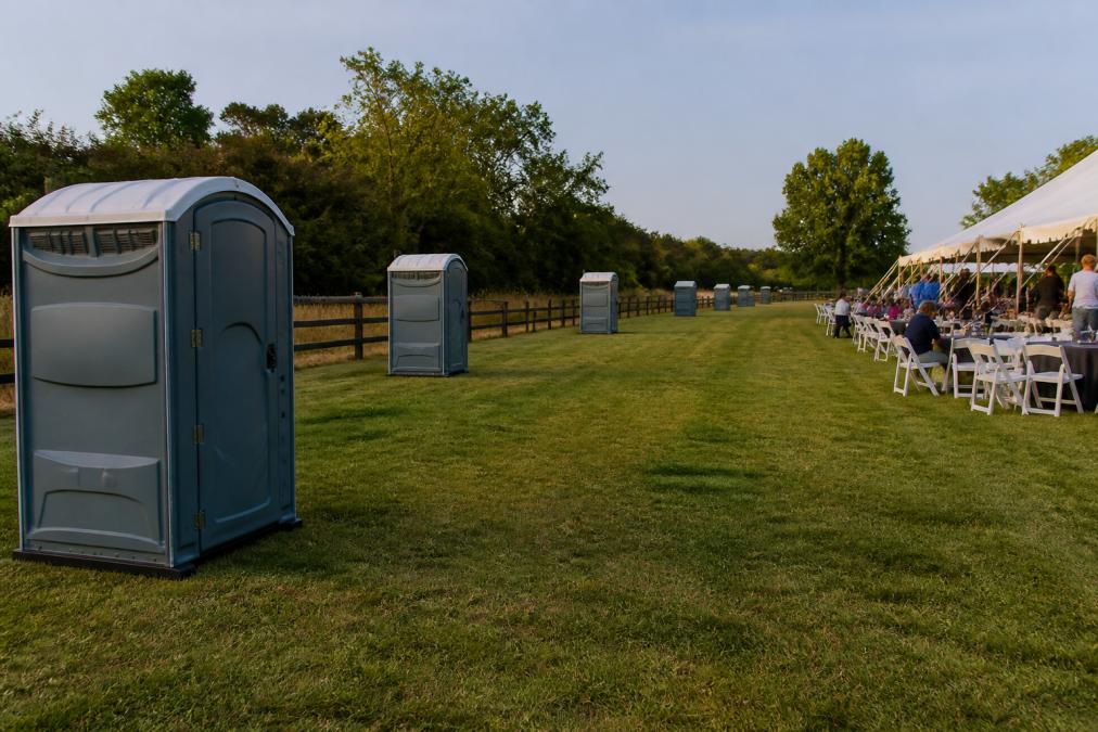 Outdoor event space with multiple portable restroom units positioned strategically at various points around the perimeter, showing proper spacing between units and clear sight lines from gathering areas, with natural daylight illuminating the setup