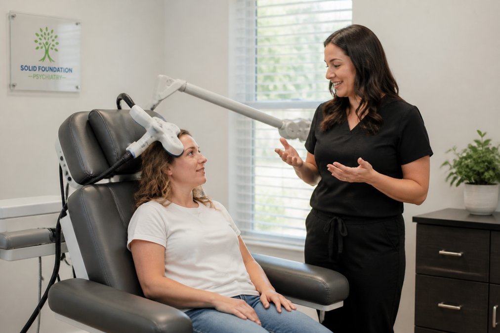 A patient seated comfortably in a modern psychiatric clinic treatment chair with a TMS device positioned near their head, a caring clinician standing nearby explaining the process, soft natural light in the room