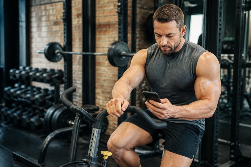A fit male bodybuilder in a gym setting, seated on a stationary bike checking his heart rate on a phone, with dumbbells and a barbell rack visible in the background under natural gym lighting