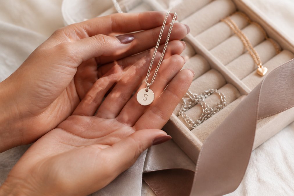 A close-up of a woman's hands holding a personalized silver necklace with an engraved initial, placed on a velvet jewelry tray with soft natural light, conveying a sense of cherished memories and sentimental value