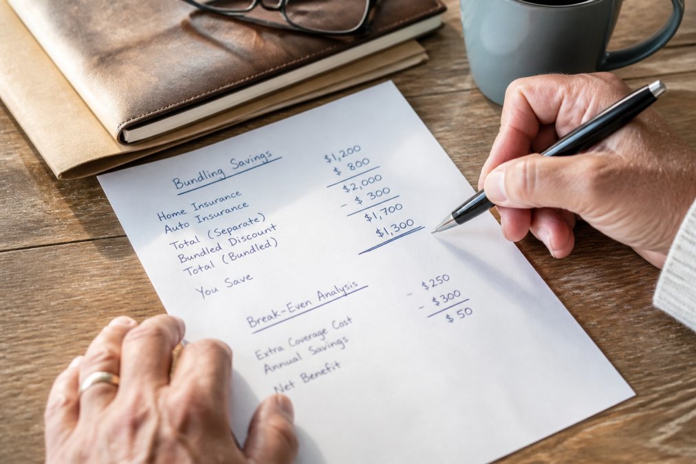 Close-up of hands holding a pen over a financial worksheet with bundling savings calculations and premium comparisons written out, natural desk lighting