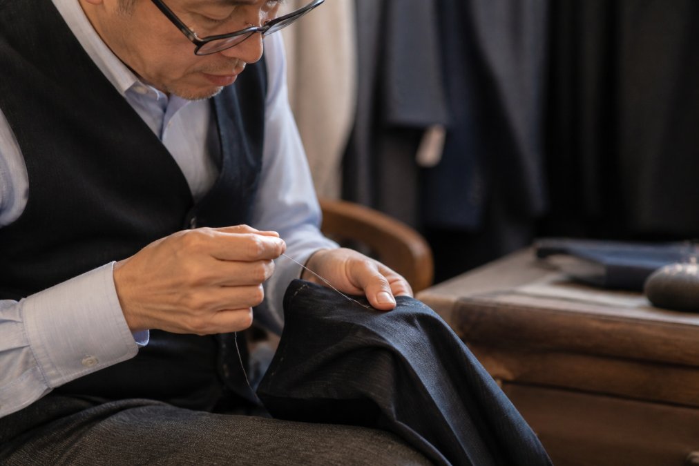 Close-up of a tailor's hands carefully hand-stitching a pant hem with needle and thread, even stitches visible on dark wool fabric under bright workbench lighting