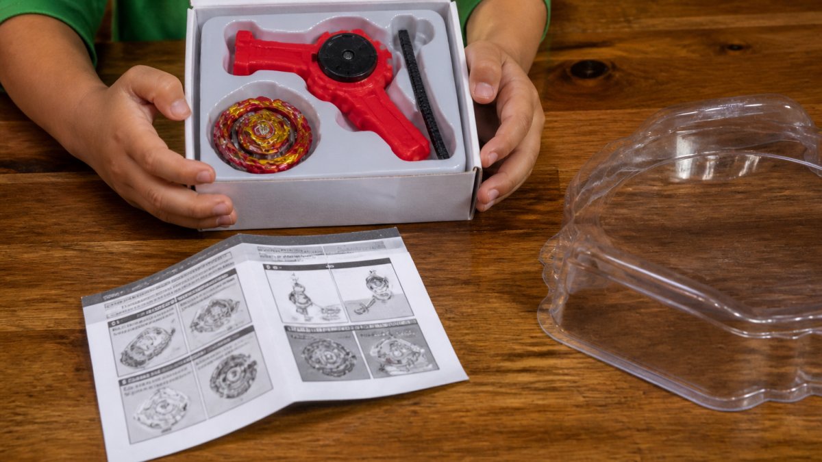 Child's hands holding an opened Beyblade starter pack, showing the spinning top, launcher, and instruction booklet laid out on a wooden table