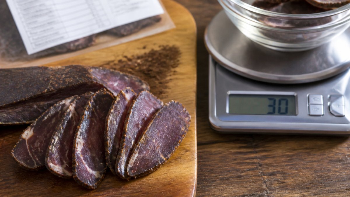 Close-up of sliced biltong pieces on a wooden board beside a small kitchen scale displaying 30g, with a nutrition label partially visible in the background