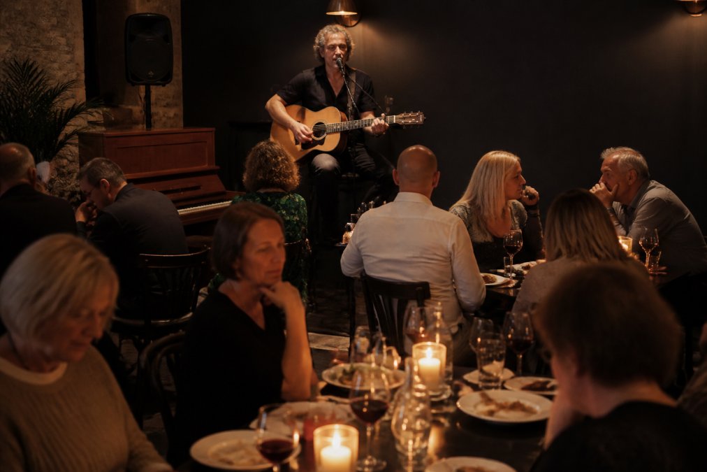 A solo acoustic guitarist performing on a small stage in a warmly lit, intimate bistro dining room, with guests at candlelit tables enjoying their meals and drinks in the background