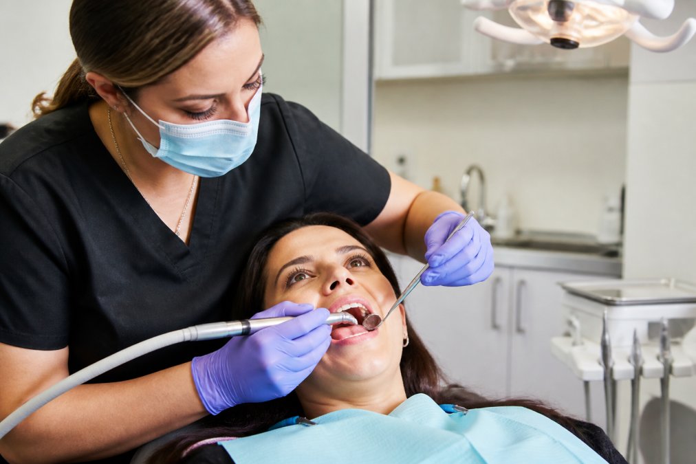 Dental hygienist performing professional cleaning on patient in modern dental office with bright overhead lighting and dental equipment visible, patient reclined in chair
