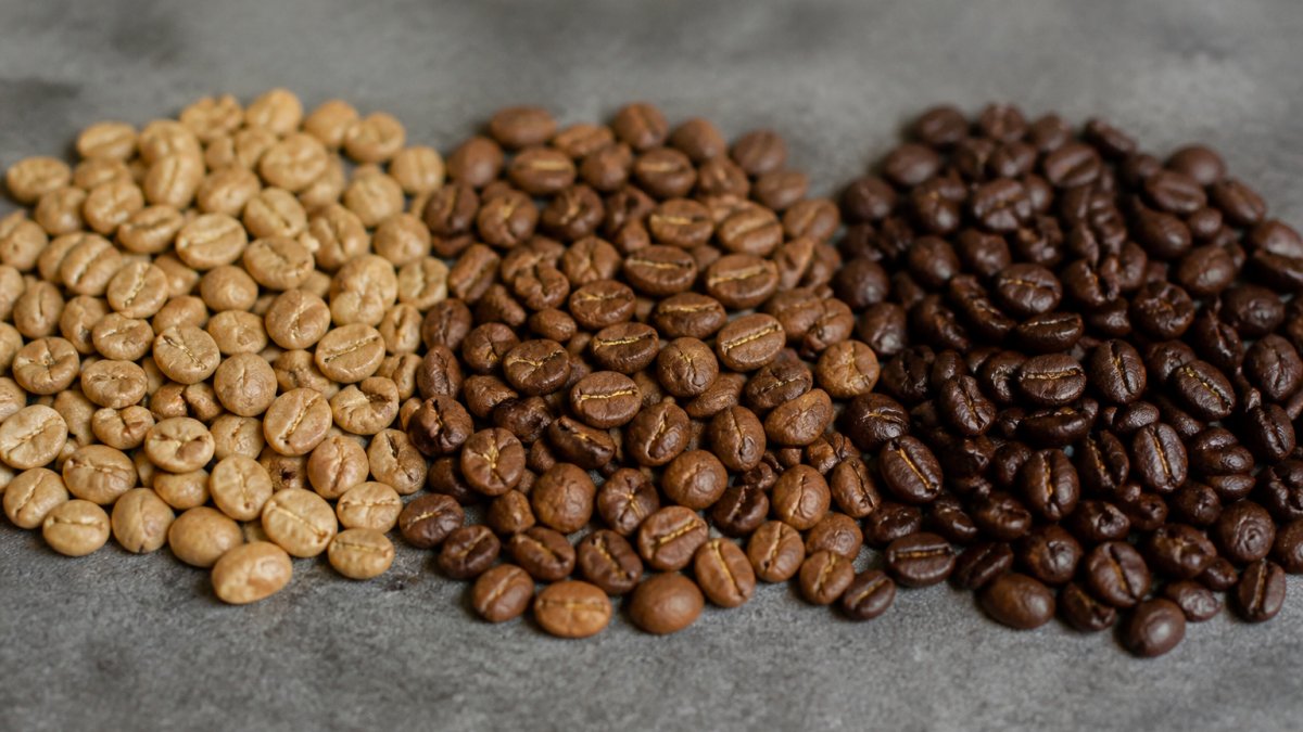Close-up of coffee beans at different roast stages, from light tan to medium brown to dark chocolate brown, arranged in a row showing visual progression of roasting development