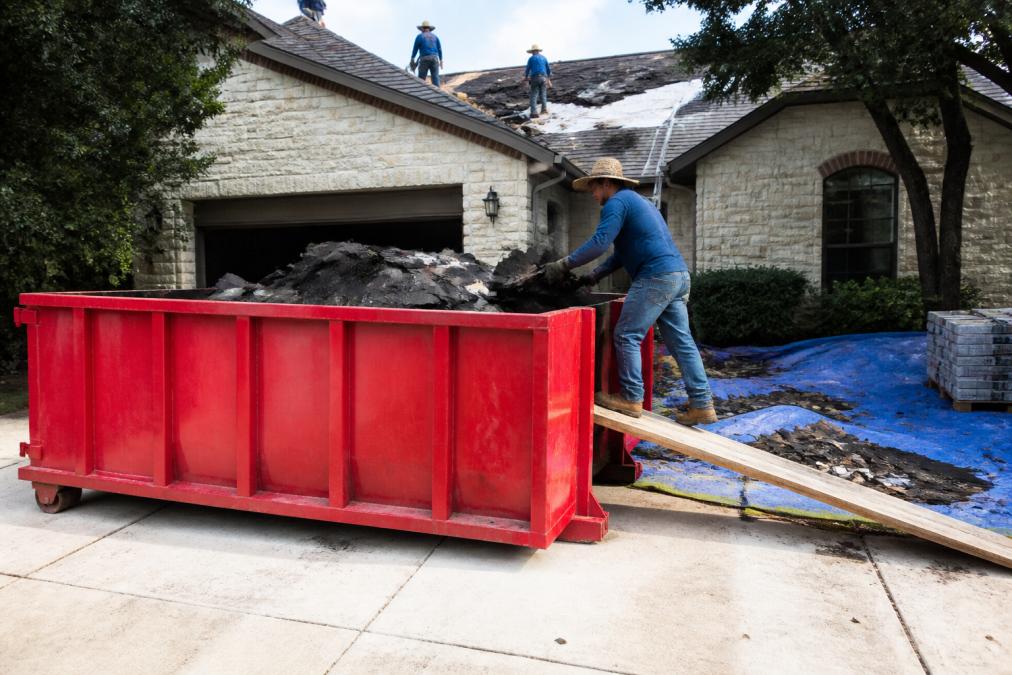 Roofing crew loading shingles and underlayment into a roll-off dumpster at a residential job site, showing organized debris management in action