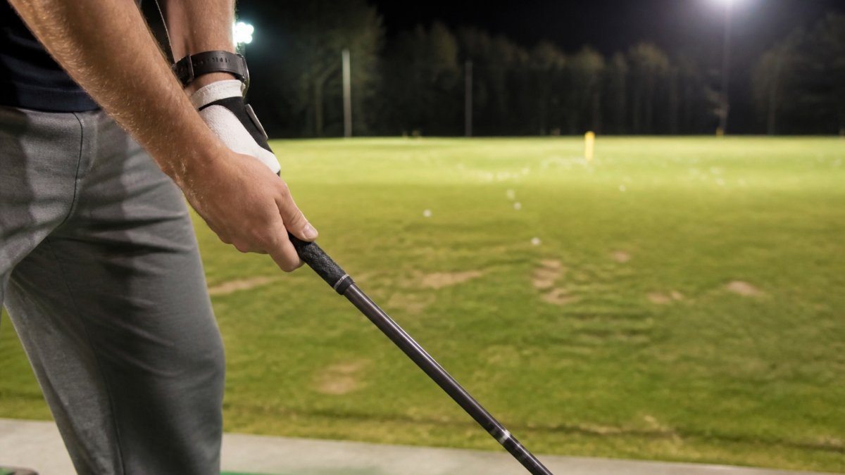 Close-up of a golfer's hands gripping a graphite driver shaft at address on a floodlit driving range, with a launch monitor screen softly blurred in the background