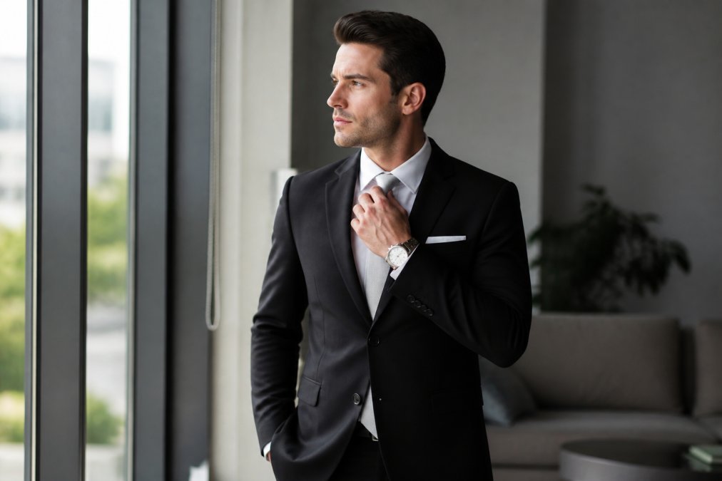 A well-dressed man in a fitted black suit stands in a bright modern room, adjusting his silver silk tie; a crisp white pocket square and minimalist silver watch are visible, with soft natural window light casting a confident silhouette