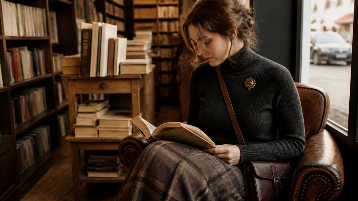 A young adult in a dark academia reading outfit, wearing a charcoal turtleneck, long plaid skirt, and vintage gold brooch, sitting in a cozy bookstore corner with a leather crossbody bag and an open book on their lap