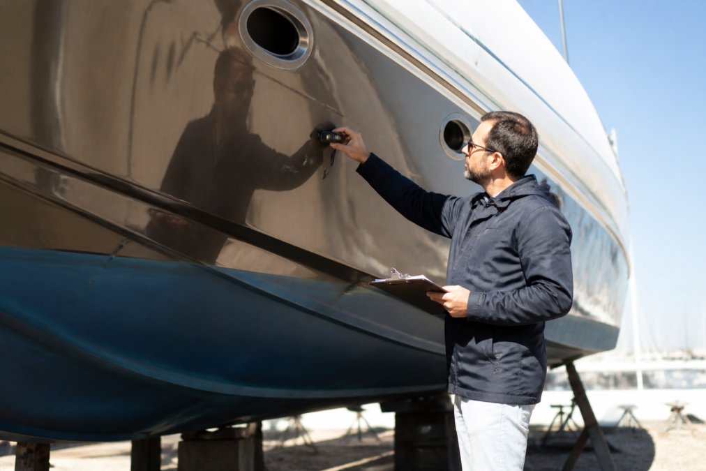 Professional yacht surveyor conducting detailed inspection of yacht hull exterior with measurement tools and clipboard, examining the vessel's condition in bright daylight