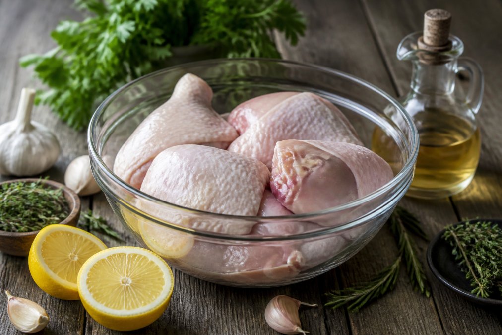Close-up of raw chicken pieces in a glass bowl surrounded by fresh lemon halves, garlic cloves, sprigs of fresh herbs and a small jug of vinegar on a rustic wooden kitchen surface