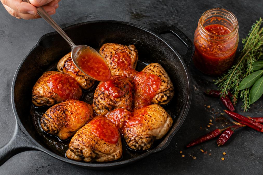 Overhead shot of a cast-iron pan with golden-brown cooked chicken being spooned over with a glossy, deep-red chilli glaze, with a small jar of artisanal hot sauce and fresh herbs beside it on a dark slate surface